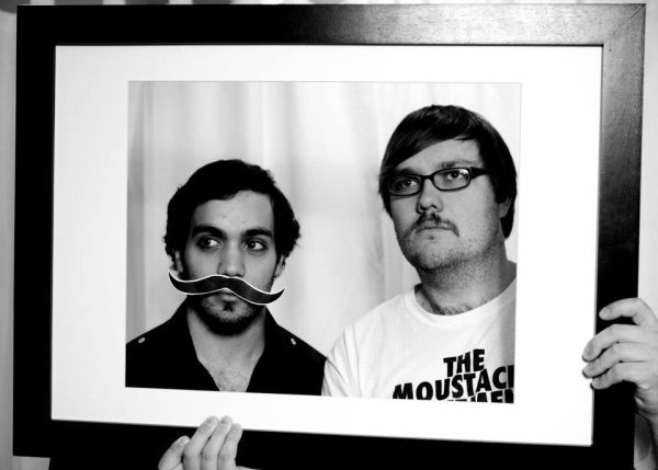 Two young men pose, one with a real mustache and the other with a fake mustache.